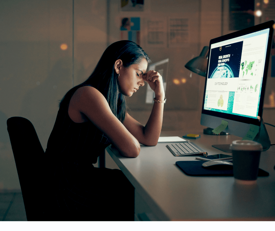 Woman at desk in a dimly lit room, illuminated by the soft glow of her computer screen. She appears overwhelmed and fatigued, as indicated by her closed eyes and a hand gently pressing against the bridge of her nose, suggesting she is experiencing burnout and stress.