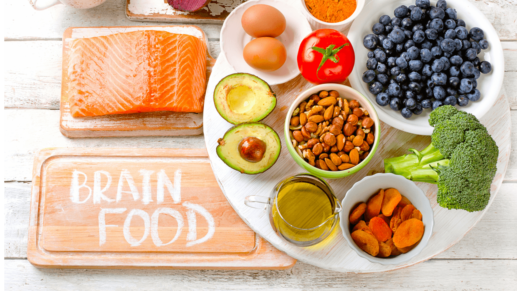 A vibrant spread of brain-healthy foods including salmon, avocados, berries, nuts, olive oil and broccoli arranged around a chalkboard reading 'BRAIN FOOD'.