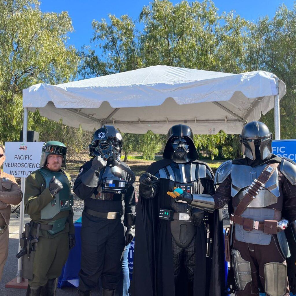 The Pacific Neuroscience Institute's booth at Walk MS Los Angeles is featured in the photograph. Four Star Wars characters, dressed in costume, stand in front of the booth.