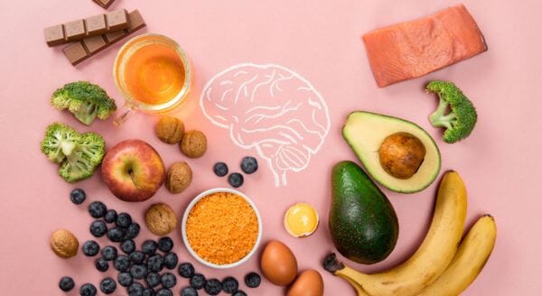Flat lay of brain-healthy foods including salmon, avocado, bananas, broccoli, blueberries, walnuts, eggs, and dark chocolate on a pink background with a white brain outline in the center.